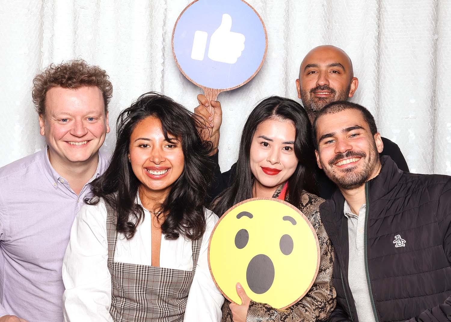 Group of five smiling individuals posing in a photo booth, holding fun props including a large surprised face emoji and a thumbs-up sign, set against a shimmering backdrop, capturing a lively and expressive moment.