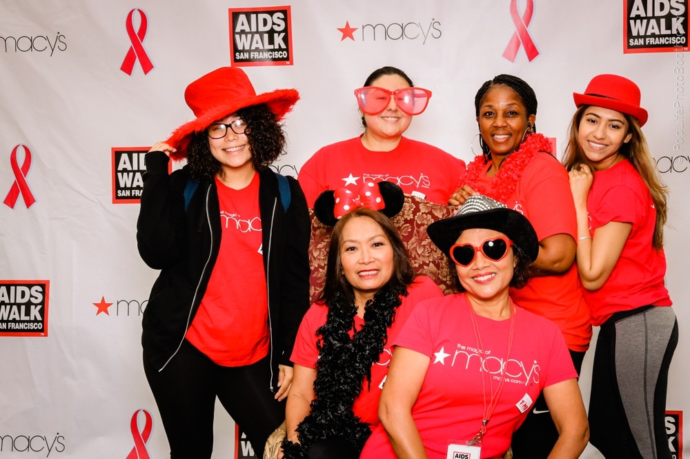 Group of participants in red shirts and fun accessories posing at the San Francisco AIDS Walk photo booth, featuring a backdrop with event branding and pink ribbons.
