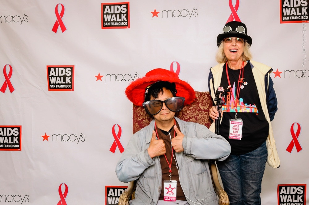 Participants at the San Francisco AIDS Walk posing in a photo booth, wearing fun costumes and accessories, with a backdrop featuring event logos and pink ribbons.