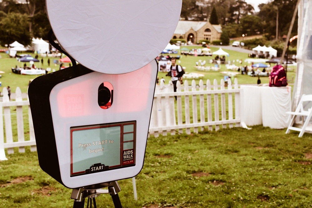 Open-air photo booth at the San Francisco AIDS Walk, featuring a screen that says "Press START to begin!" with event details visible in the background, capturing the festive atmosphere of the fundraising event.