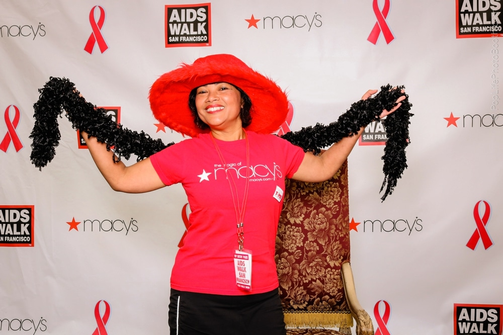 Smiling participant in a red hat and matching shirt at the San Francisco AIDS Walk, posing with a feather boa in front of a branded backdrop featuring event logos and pink ribbons.