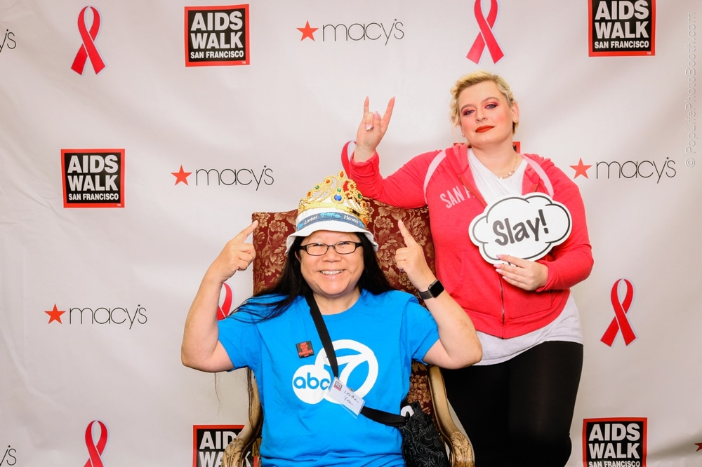 Participants at the San Francisco AIDS Walk posing in a photo booth, one wearing a crown and holding a sign saying "Slay!" against a backdrop featuring event branding and sponsor logos.