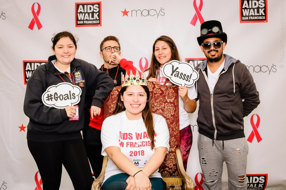 Group of participants at the San Francisco AIDS Walk posing in a photo booth, holding signs with "#Goals" and "Yasss!", wearing event shirts and festive attire, with a backdrop featuring the event logo and sponsor Macy's.