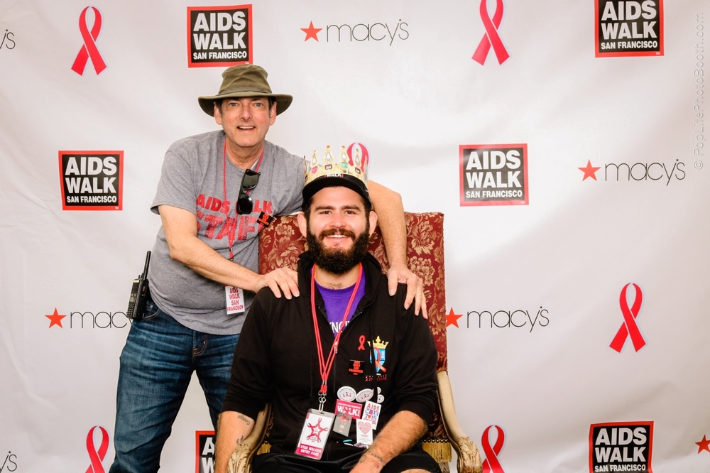 Two participants at the San Francisco AIDS Walk posing in front of a branded backdrop, one wearing a crown and both displaying team spirit with event badges.