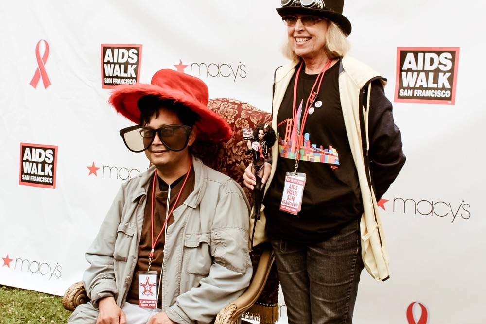 Participants at the San Francisco AIDS Walk posing for a photo booth, featuring a man in oversized glasses and a red hat, alongside a woman in a black shirt with a colorful design, both smiling against a branded backdrop.