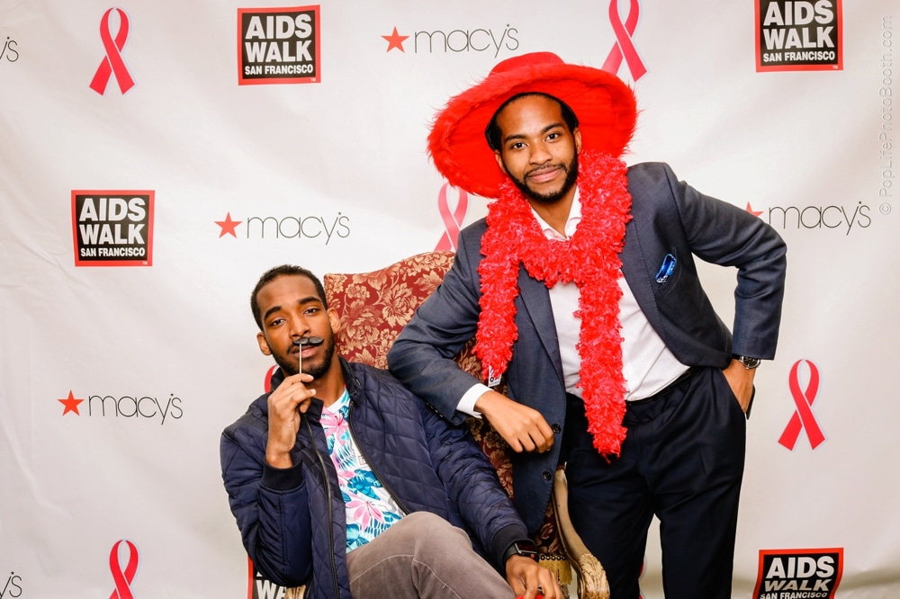 Two men posing with playful expressions at the San Francisco AIDS Walk photo booth, one wearing a red feather boa and hat, with a backdrop featuring the event's logo and Macy's branding.