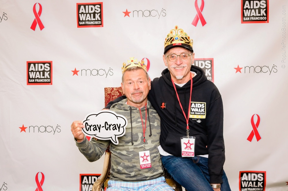 Two men wearing crowns and "AIDS Walk" shirts posing together in front of a backdrop featuring the AIDS Walk logo and Macy's branding, holding a speech bubble sign that says "Cray-Cray."