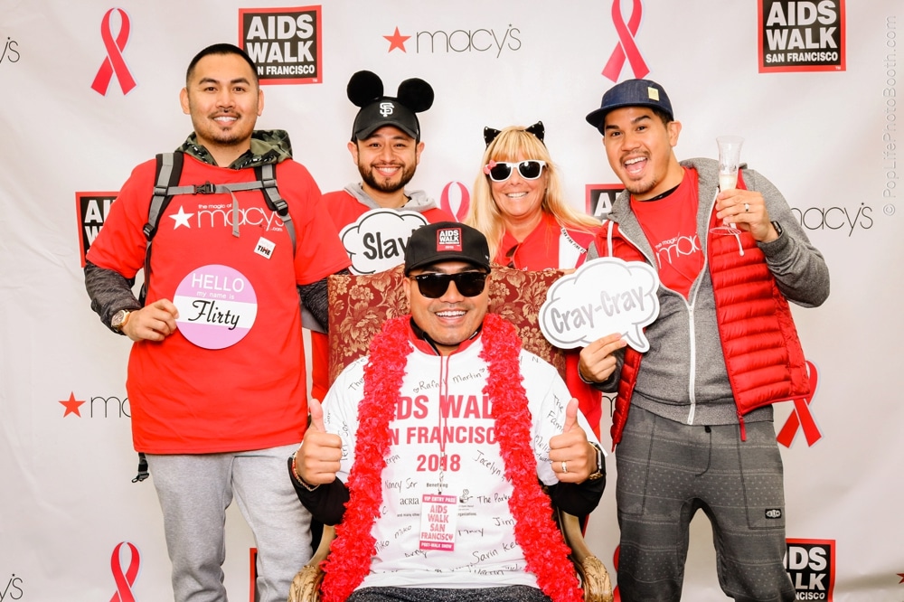 Group of participants at the San Francisco AIDS Walk, wearing themed shirts and accessories, posing with playful signs in front of a branded backdrop featuring the event's logo and Macy's.
