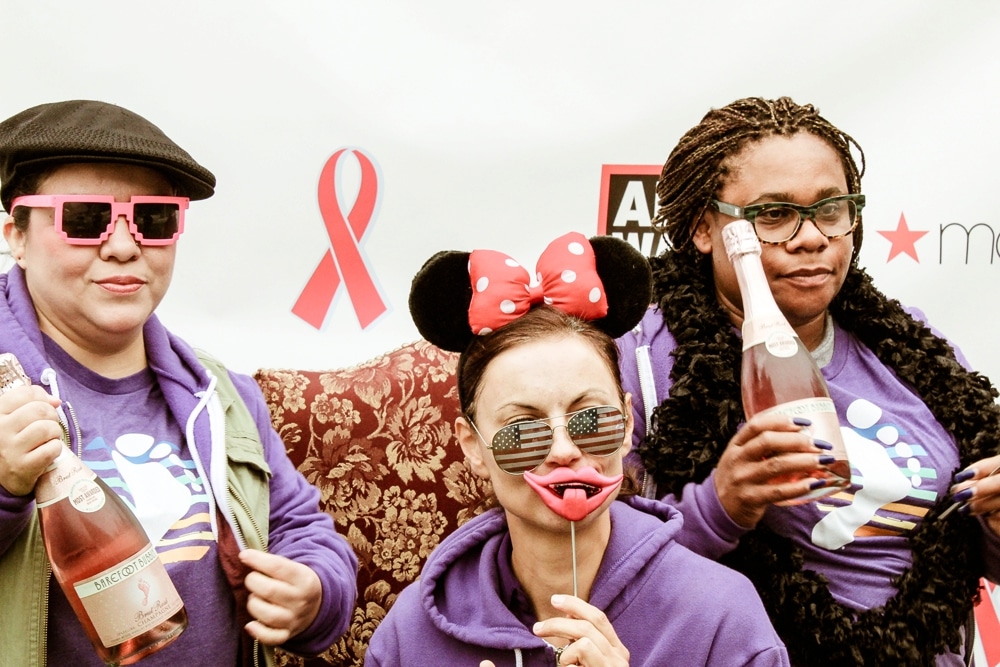 Three participants in vibrant costumes celebrating at the San Francisco AIDS Walk photo booth, featuring playful props like oversized sunglasses, a Minnie Mouse headband, and a novelty lip prop, against a backdrop with event branding.