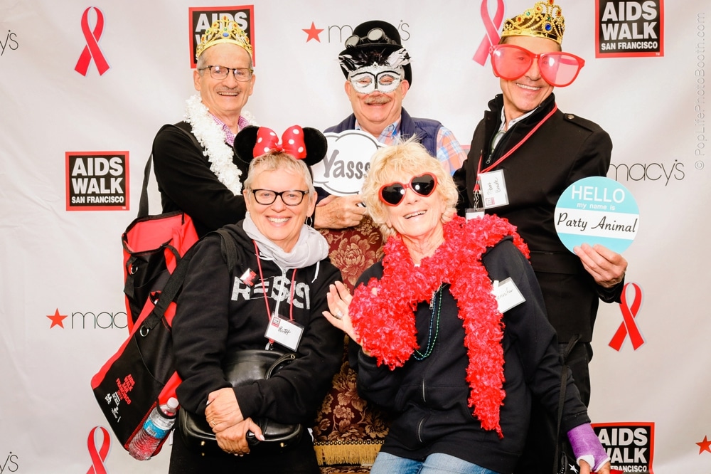 Group of participants at the San Francisco AIDS Walk posing in a photo booth, wearing colorful costumes and accessories, with signs reading "Yasss" and "Hello, my name is Party Animal," against a backdrop featuring event branding.