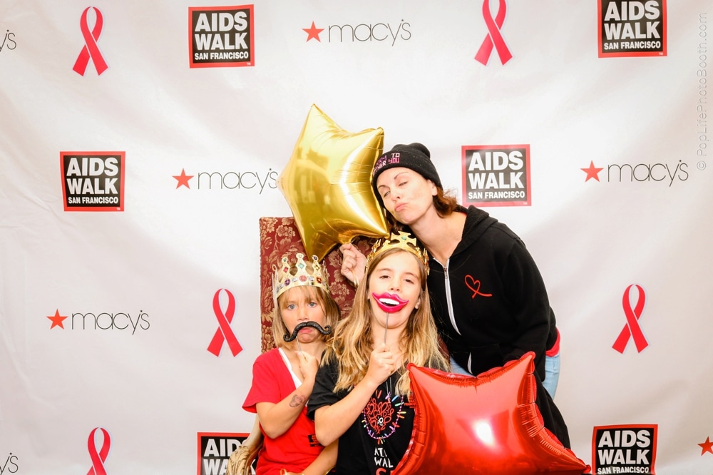 Three participants at the San Francisco AIDS Walk posing in a photo booth, wearing fun accessories like a crown and a mustache, holding balloons, with branded backdrop featuring AIDS Walk and Macy's logos.