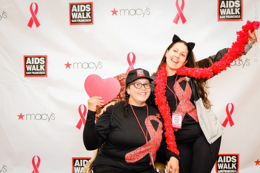 Two women posing at the AIDS Walk San Francisco photo booth, wearing matching black shirts with red ribbon designs, holding a heart prop, decorated with a red feather boa, against a backdrop featuring AIDS Walk and Macy's logos.