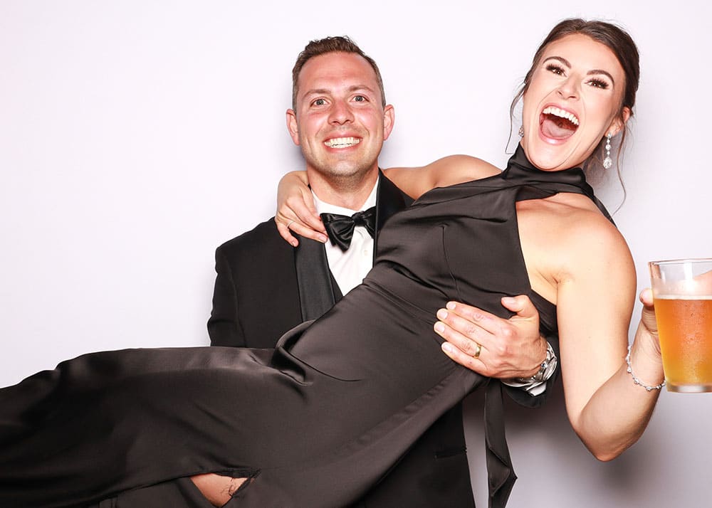 Couple posing in a playful pick-up pose, man in tuxedo lifting woman in a black dress, both smiling joyfully, with a drink in her hand, against a plain background, capturing a fun photo booth moment.