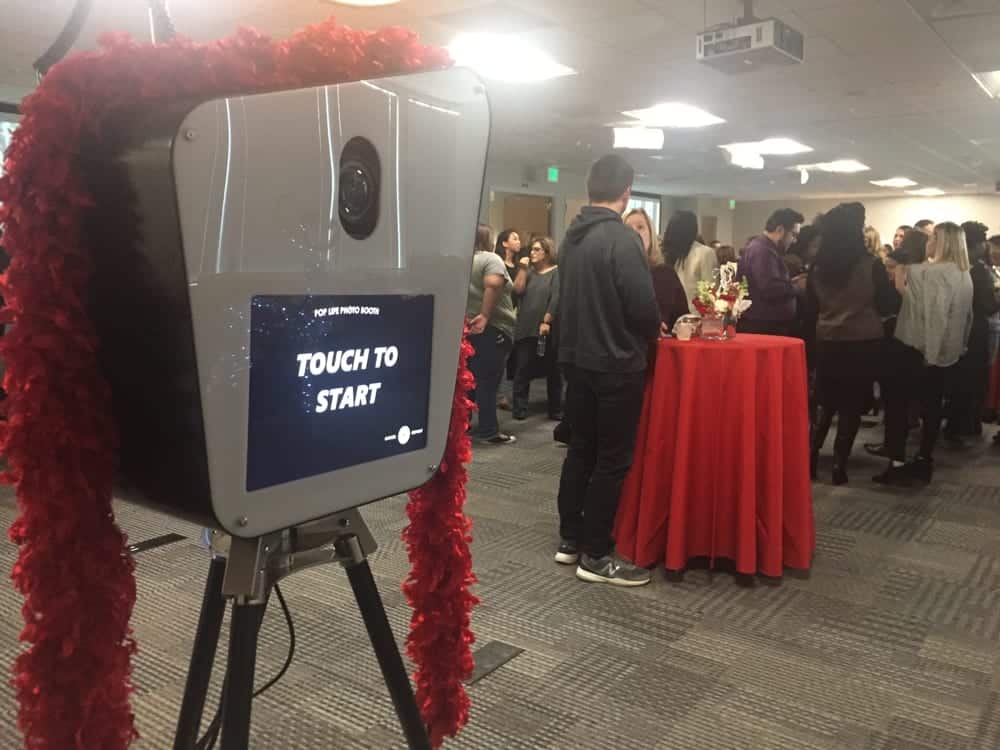 Photo booth with "Touch to Start" screen and decorative red accents, set up at a lively event with guests mingling in the background.