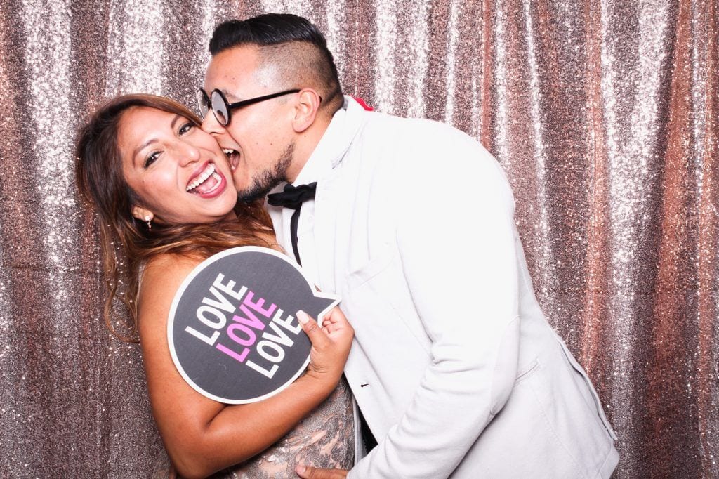 Couple joyfully posing in a photo booth, holding a sign that reads "LOVE LOVE LOVE," with a sparkling backdrop, capturing the fun and excitement of a wedding celebration.