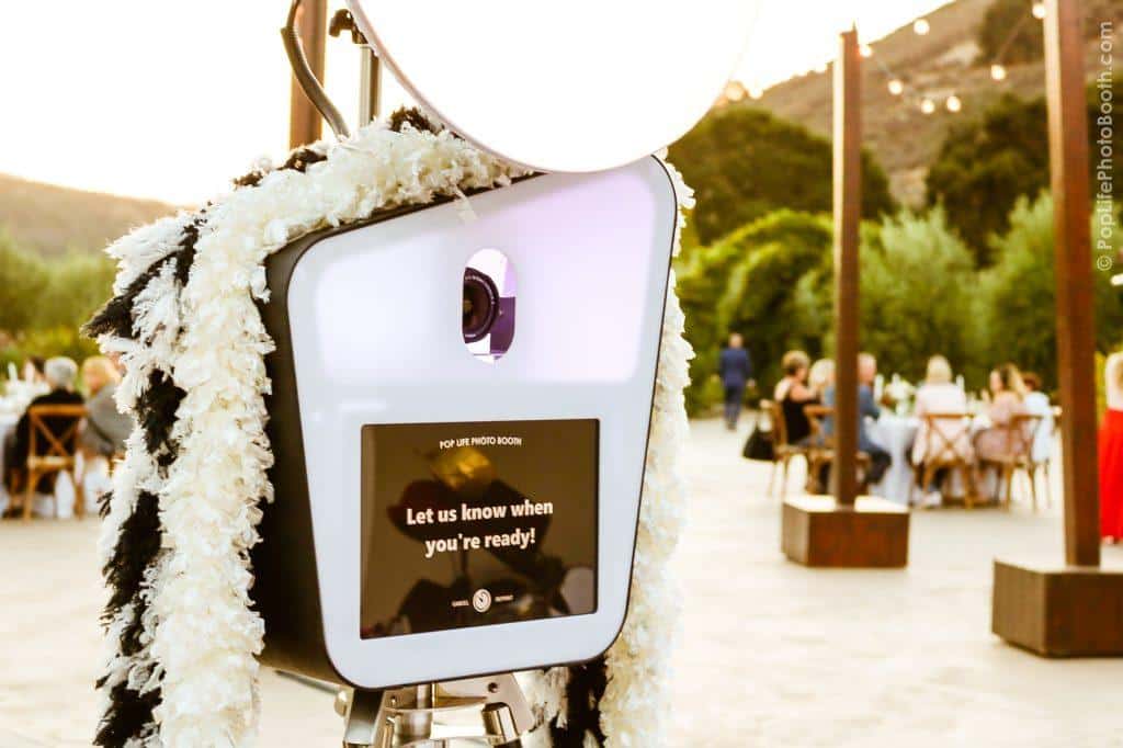 Pop Life Photo Booth setup with a digital screen displaying "Let us know when you're ready," surrounded by guests enjoying a wedding reception in an outdoor setting.