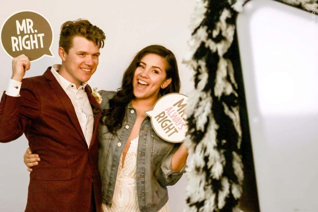Couple posing in a photo booth, holding signs "MR. RIGHT" and "MRS. ALWAYS RIGHT," celebrating their wedding with fun props and smiles.