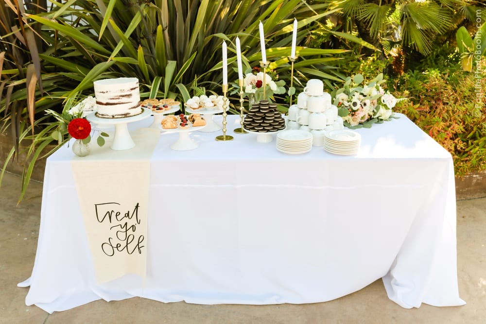 Dessert table featuring a multi-layered cake, assorted pastries, and a "Treat Yo Self" banner, surrounded by greenery, enhancing the festive atmosphere of the wedding celebration for Thomas and Anastasha.