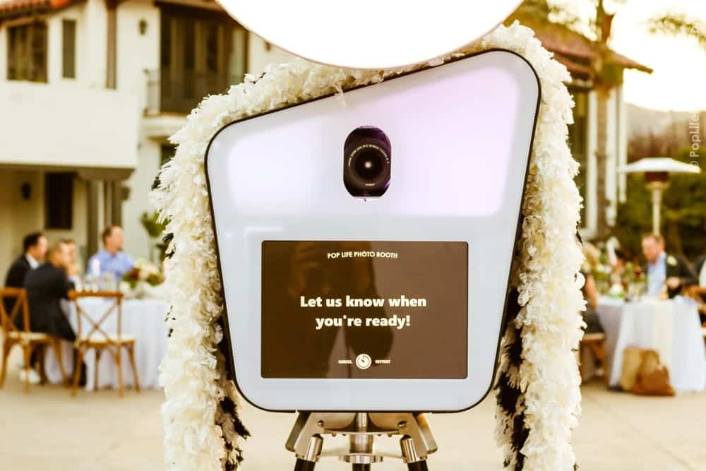 Pop Life Photo Booth setup with floral decorations and a screen displaying "Let us know when you're ready!" at a wedding reception, capturing the festive atmosphere of Thomas and Anastasha's celebration.