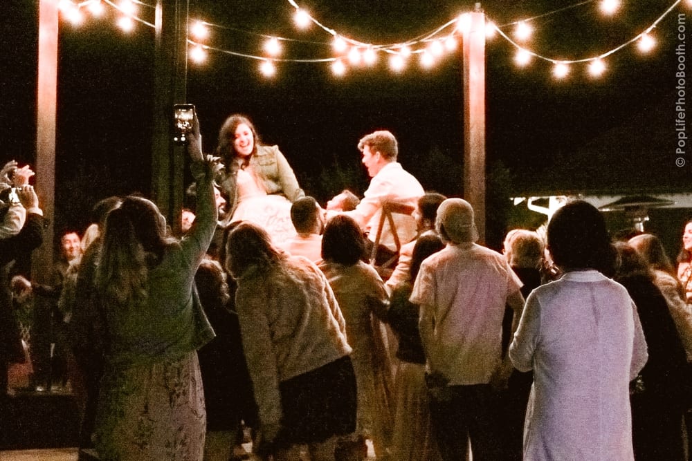 Guests joyfully participating in a celebratory chair dance at Thomas and Anastasha’s wedding reception, illuminated by string lights, showcasing the lively atmosphere and fun moments captured by Pop Life Photo Booth.