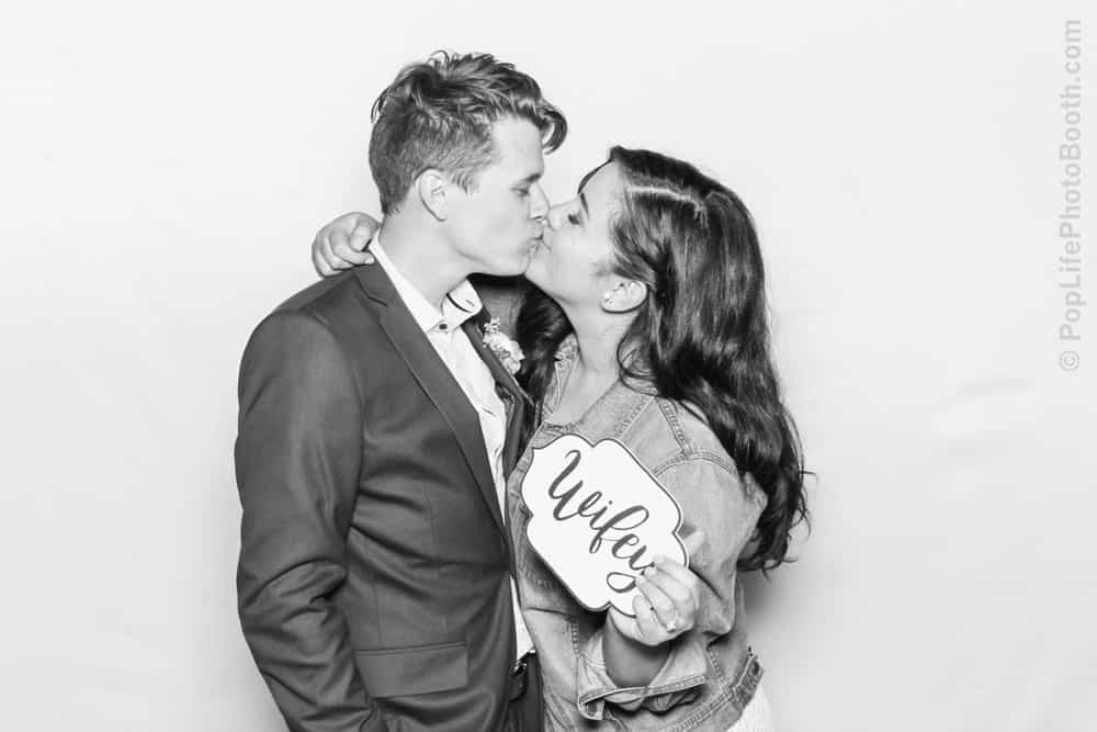 Couple kissing in photo booth, holding sign that says "Wifey," capturing joyful moment from Thomas and Anastasha's wedding celebration.