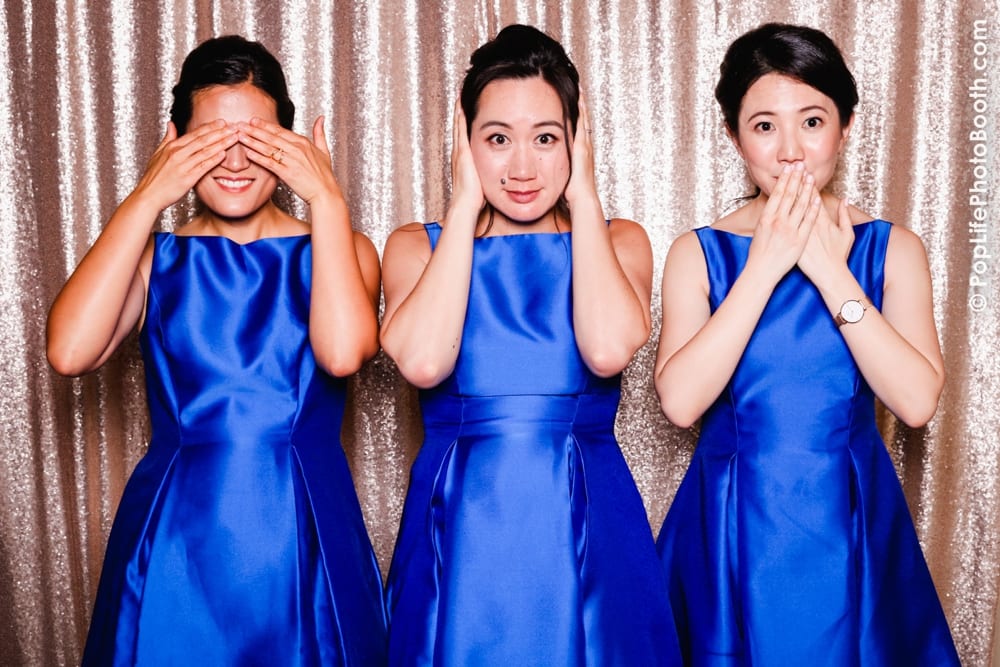 Three women in blue dresses posing in a photo booth, demonstrating the "speak no evil, see no evil, hear no evil" concept with playful expressions against a sparkling backdrop.