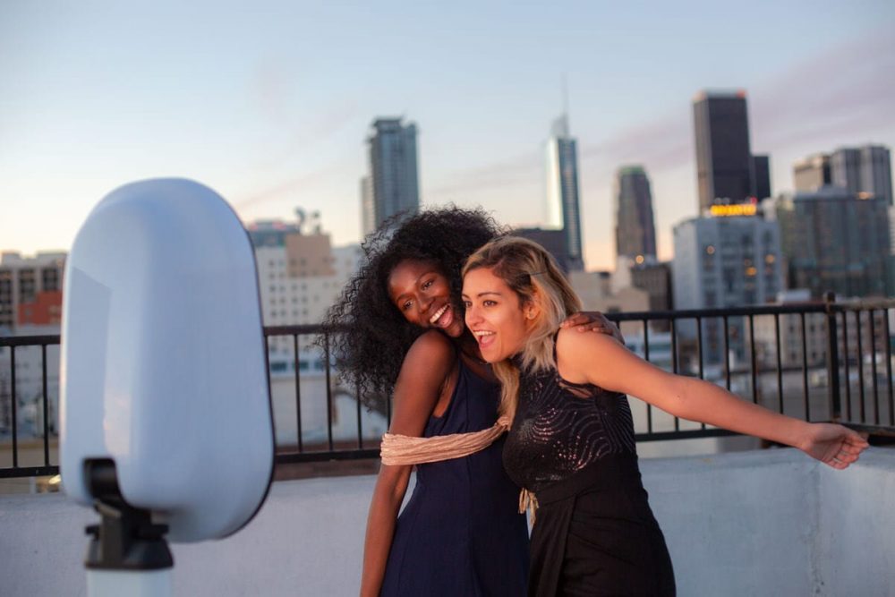Two women posing joyfully in front of a Party Booth with a city skyline backdrop, highlighting the booth's stylish design and capturing fun moments at an event.