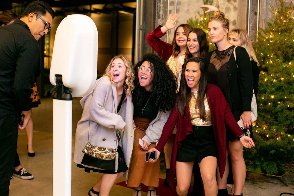 Group of excited young women posing in front of a stylish Party Booth with a white oval design, celebrating at a festive event, surrounded by decorative greenery and warm lighting.