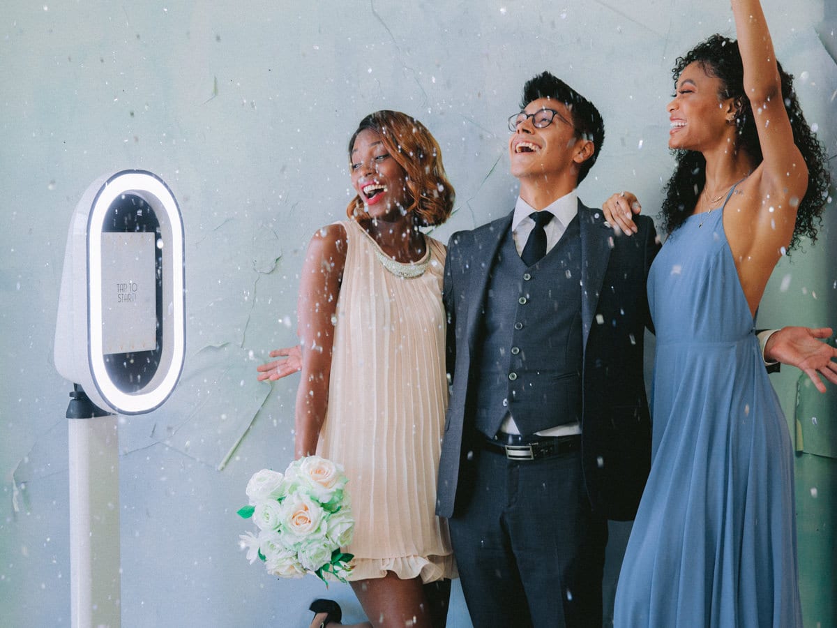 Three joyful individuals celebrating in front of a modern photo booth with a bright ring light, surrounded by falling confetti, perfect for events like weddings and parties.