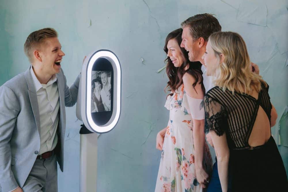 Group of four friends joyfully interacting with a modern photo booth featuring a bright oval ring light, capturing fun memories at an event, showcasing the Party Booth experience from Pop Life Photo.