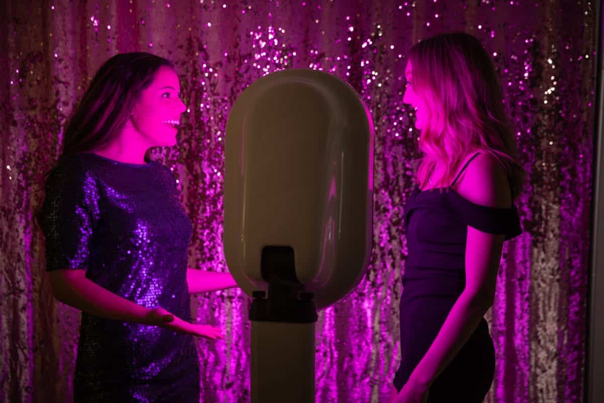 Two women smiling and interacting in front of a stylish Party Photo Booth with colorful lighting, surrounded by a shimmering pink backdrop, emphasizing fun at events like weddings and parties.