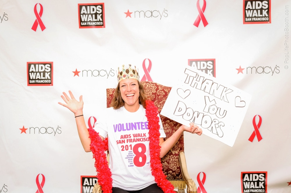 Volunteer at AIDS Walk San Francisco holding a "Thank You Donors" sign, wearing a crown and festive attire, with a backdrop featuring event branding and pink ribbons.