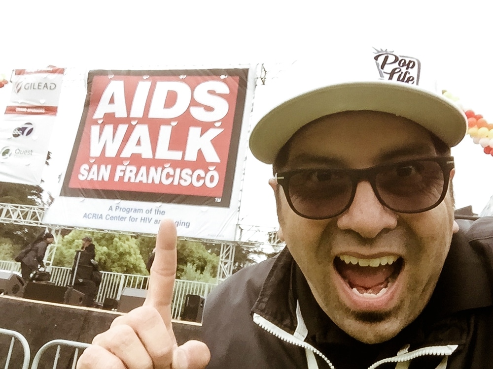 Man gesturing excitedly in front of an "AIDS Walk San Francisco" banner, promoting the fundraising event, with "Pop Life" logo on his cap.