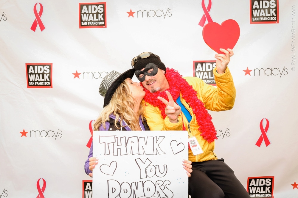 Couple posing in front of AIDS Walk San Francisco backdrop, holding a "Thank You Donors" sign, celebrating team spirit with costumes and props.