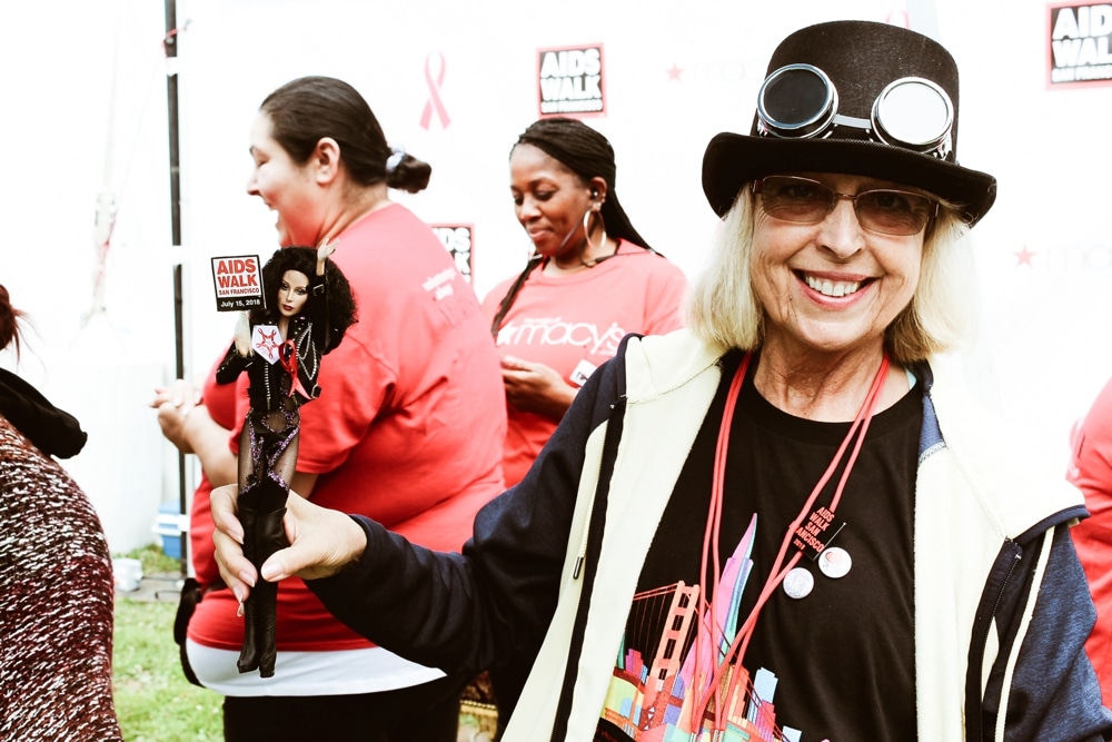 Smiling woman wearing a top hat and glasses holding a Barbie-sized Cher doll at the San Francisco AIDS Walk, with participants in red shirts and event backdrop visible.