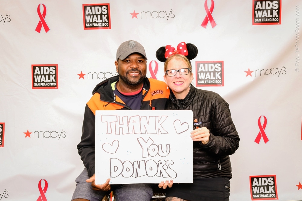 Smiling participants at the San Francisco AIDS Walk holding a sign that says "THANK YOU DONORS," with a backdrop featuring AIDS Walk branding and event sponsors.