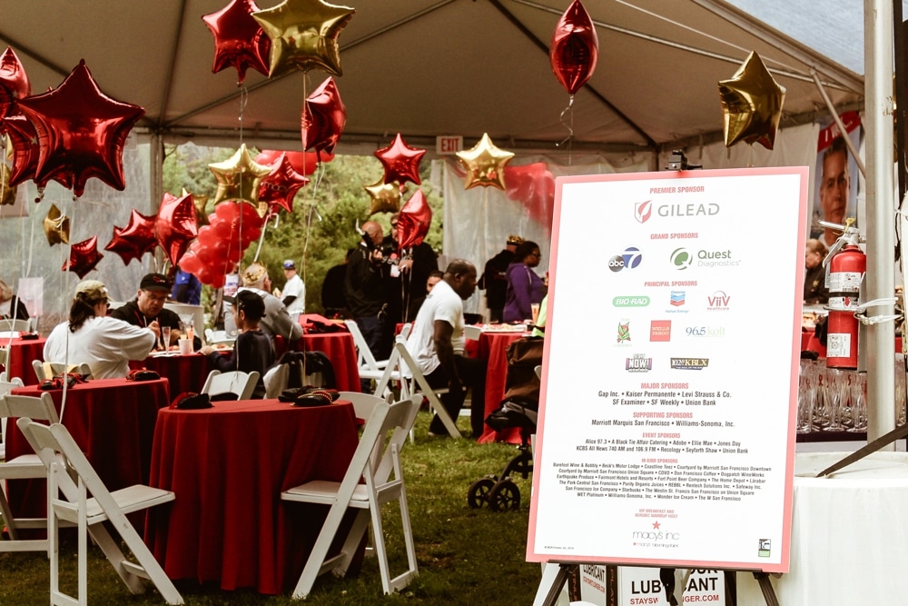 Event tent at San Francisco AIDS Walk featuring red and gold star balloons, tables with red tablecloths, and a sponsorship sign highlighting Gilead and other sponsors, capturing the festive atmosphere of the fundraising event.