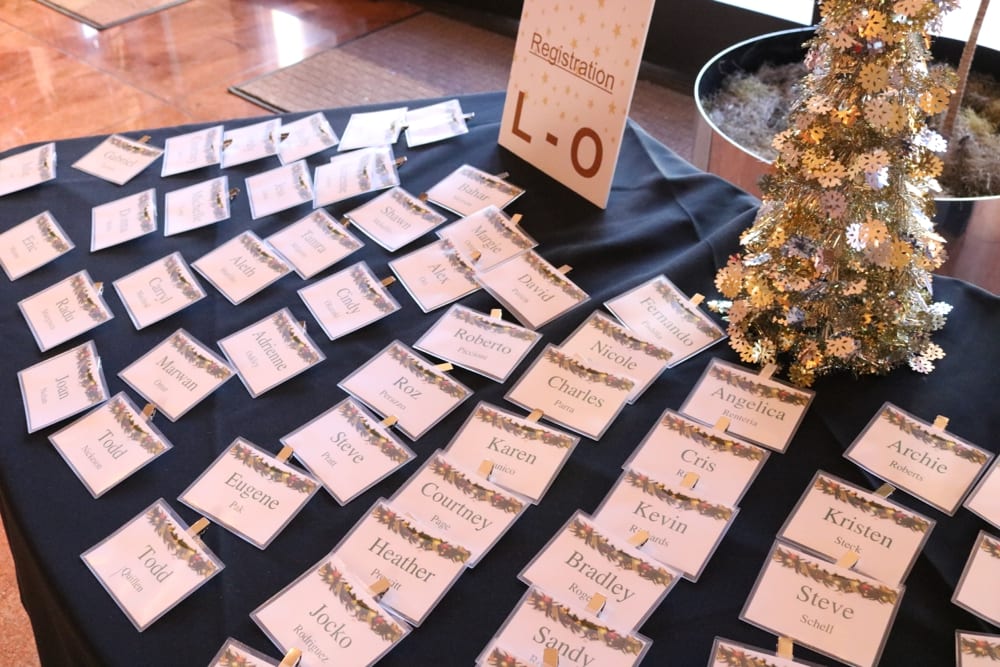 Name tags arranged on a black table with a decorative registration sign for attendees, featuring a small festive tree adorned with snowflakes and a gold star.