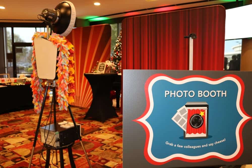 Photo booth setup with colorful decorations, featuring a sign that reads "PHOTO BOOTH" and encourages guests to "Grab a few colleagues and say cheese!"