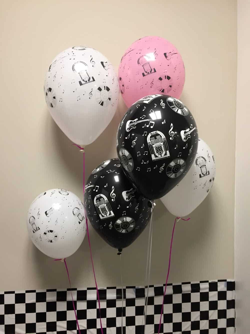 Balloons in black, white, and pink featuring music-themed designs, including jukeboxes, guitars, and musical notes, set against a checkered backdrop, enhancing event decor for celebrations and parties.