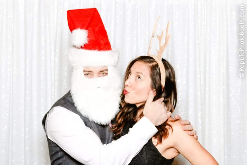 Santa Claus figure with a white beard and red hat posing with a woman wearing antlers, against a sparkling white backdrop, capturing festive holiday spirit for corporate party memories.