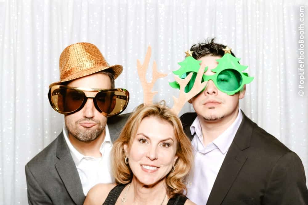 Three smiling individuals in festive attire posing in a photo booth, wearing oversized sunglasses, a glittery hat, and playful holiday-themed props against a shimmering white backdrop, capturing joyful moments for a holiday party celebration.