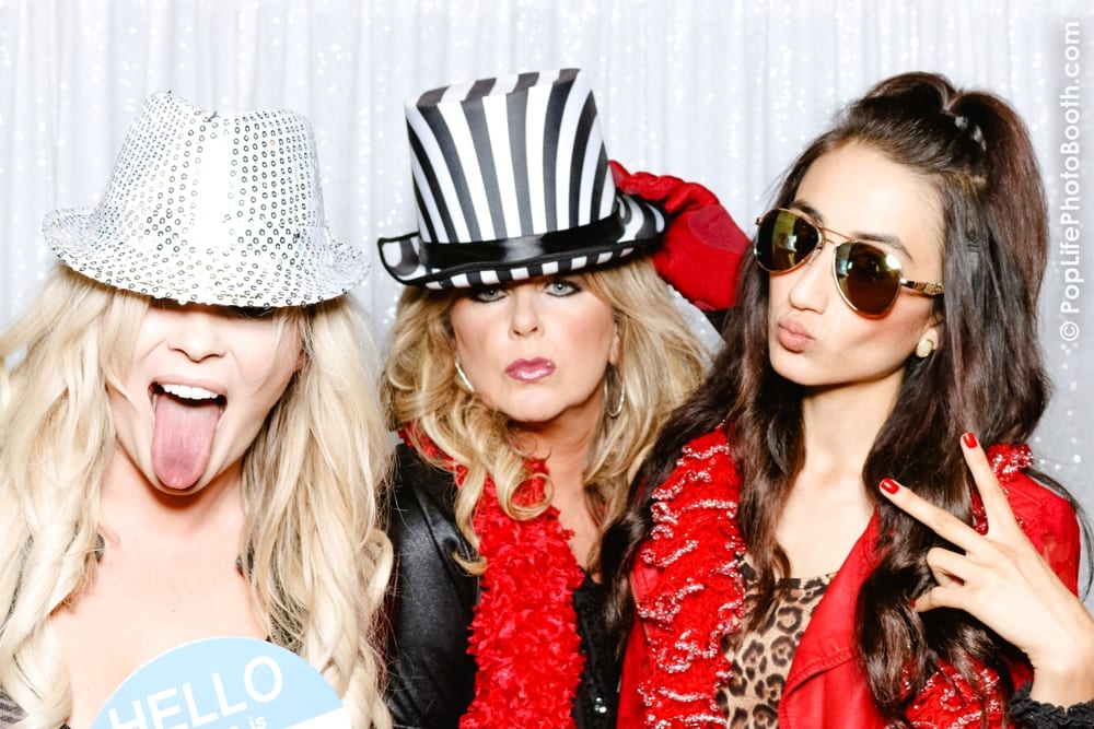 Three women posing in a photo booth with playful expressions, wearing festive hats and accessories, against a shimmering silver backdrop, capturing the fun and excitement of a holiday party celebration.