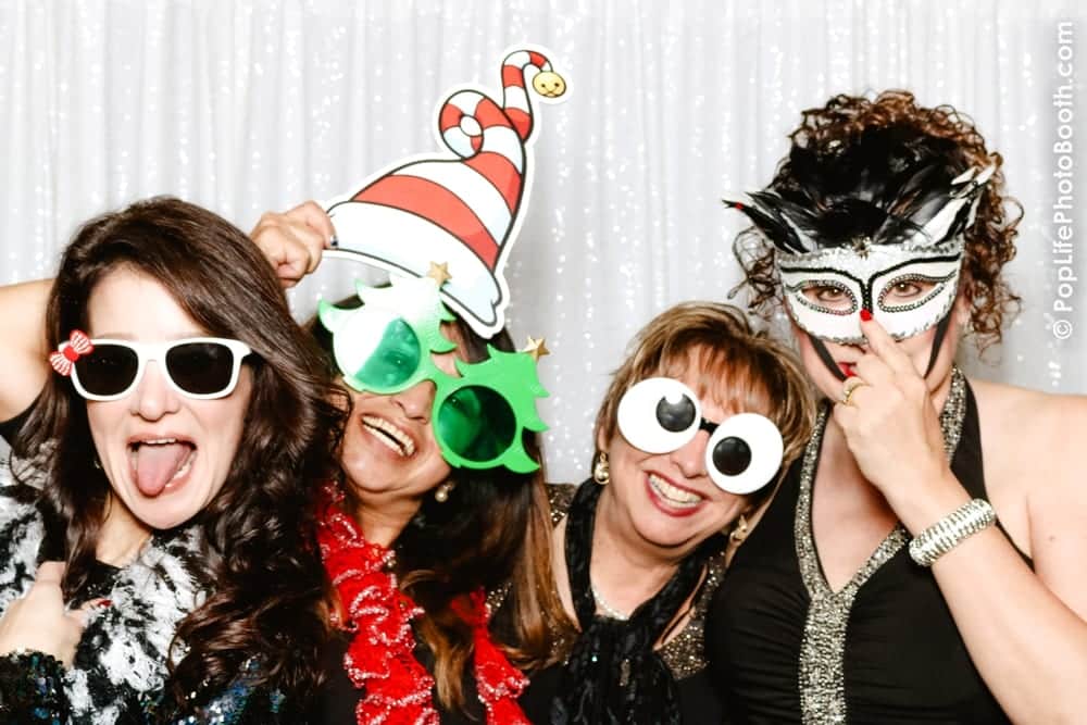 Four women posing in a photo booth with festive props, including a Santa hat, Christmas tree glasses, and a masquerade mask, against a sparkling white backdrop, capturing the fun spirit of a holiday party.