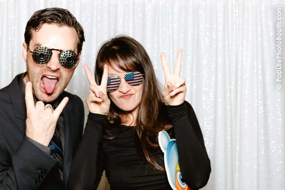 Couple posing with playful expressions and themed sunglasses in front of a shimmering backdrop, capturing a fun moment at a holiday party photo booth.