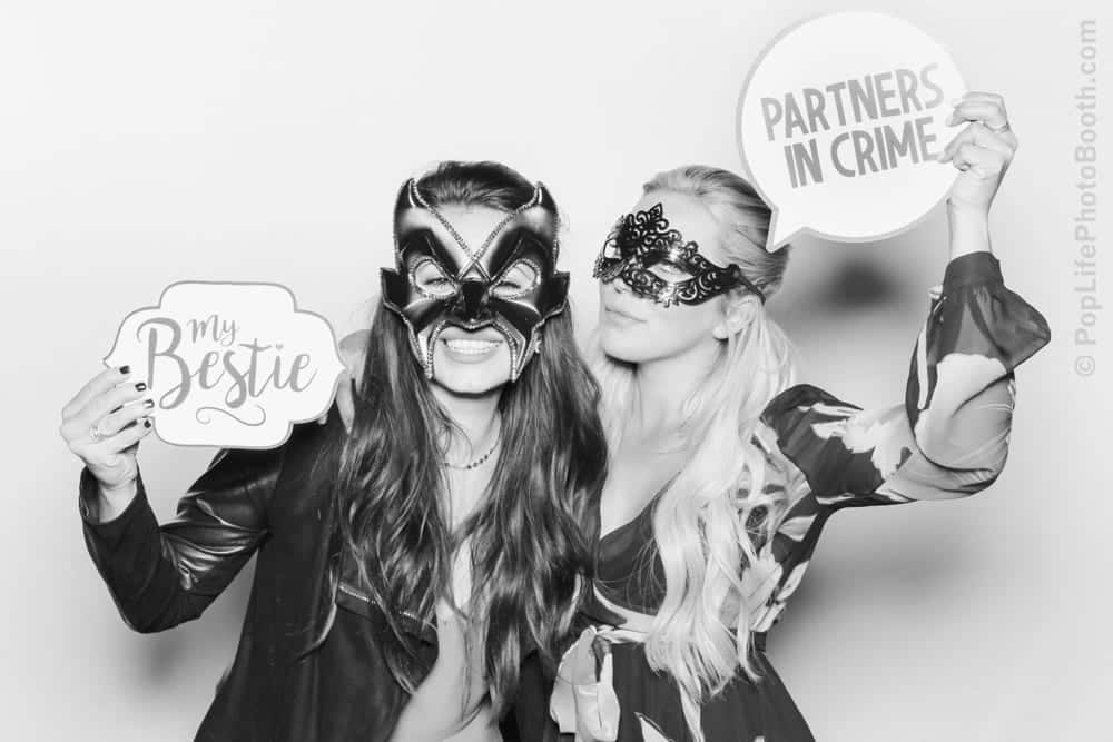 Two women wearing masks posing for a photo booth, one holding a sign that reads "My Bestie" and the other holding a sign that says "Partners in Crime," against a simple white backdrop, perfect for a masquerade ball theme.