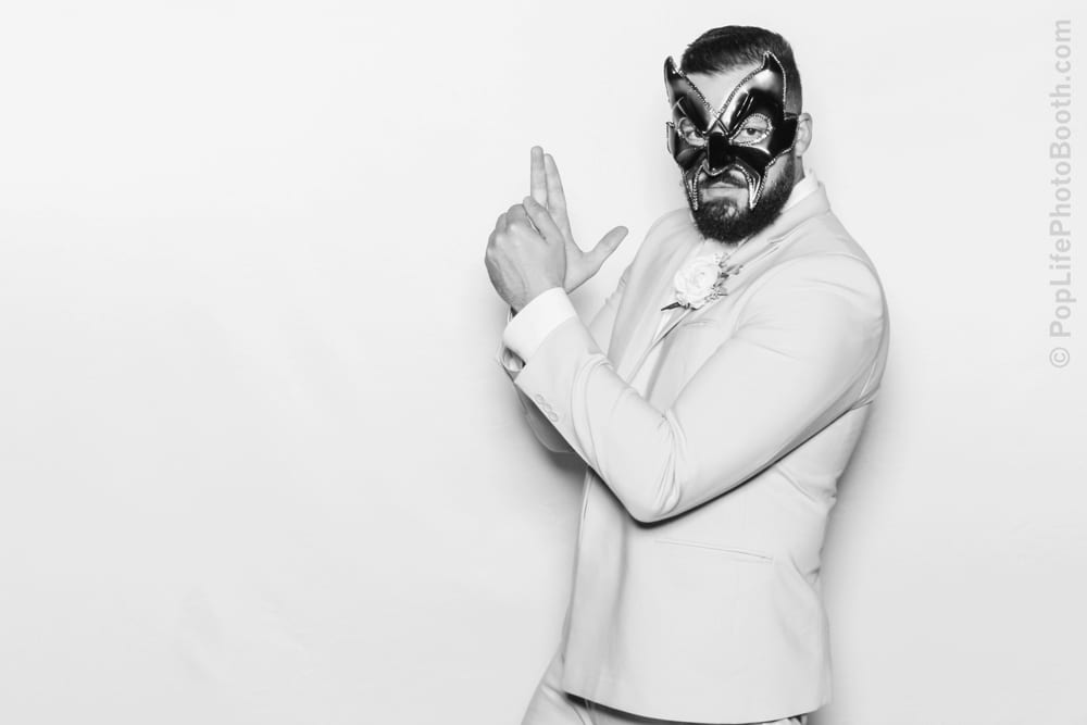 Man in white suit and black masquerade mask posing playfully against a white backdrop, embodying a festive spirit for a masquerade ball-themed event.