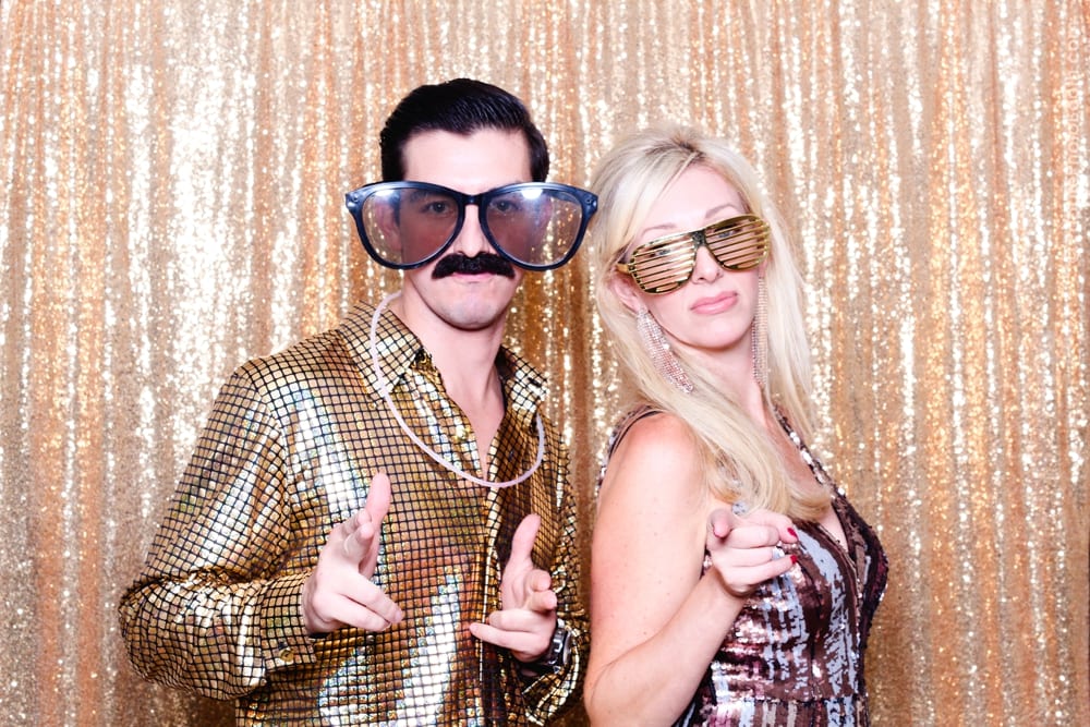 Man in shiny gold shirt and oversized glasses, woman in sparkly dress, posing playfully against a gold sequined backdrop, capturing a festive Studio 54-themed party vibe.