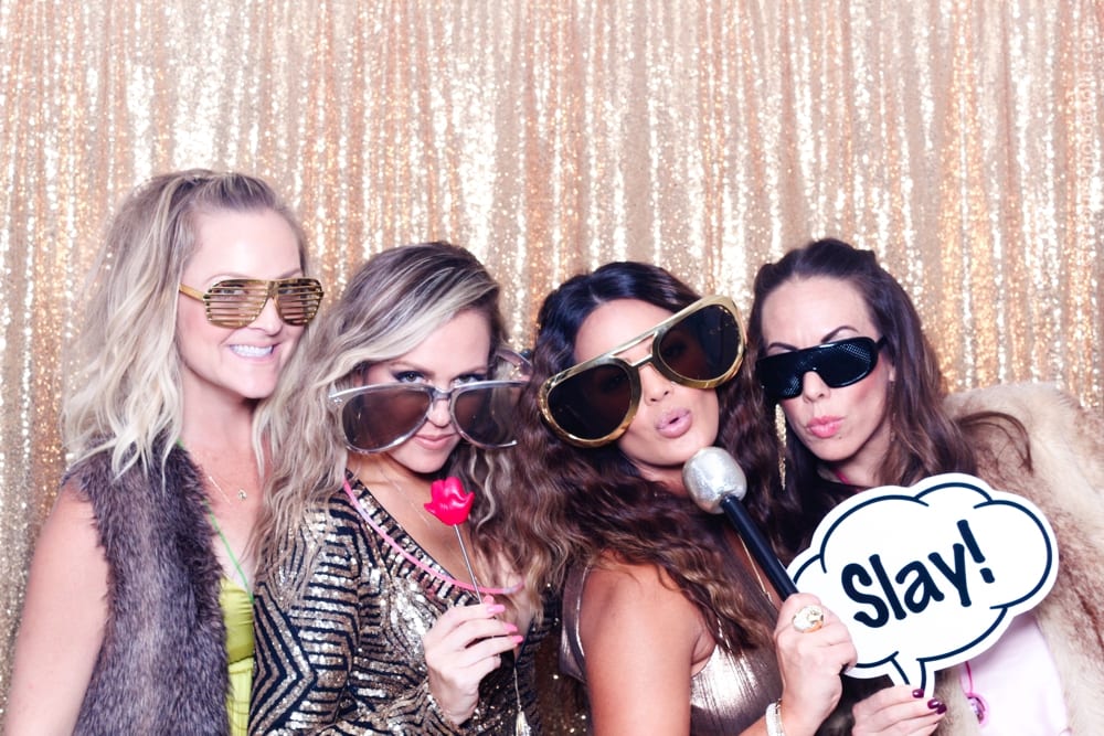 Four women posing in oversized sunglasses and glamorous outfits, holding a microphone and a sign that says "Slay!" in front of a shimmering gold backdrop, capturing a fun moment at a themed party photo booth.