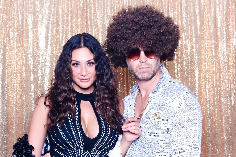 Couple posing in retro 70s attire with a gold sequined backdrop, showcasing a fun photo booth experience for a Studio 54-themed party.